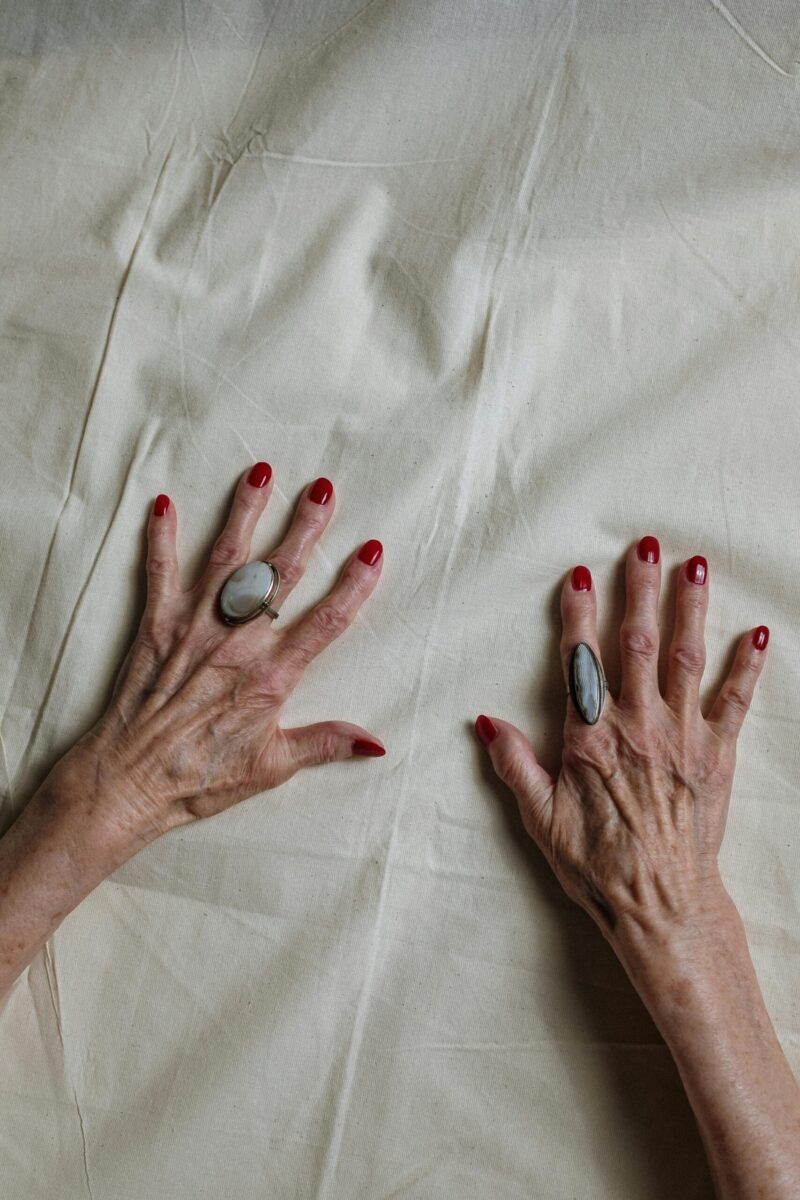 Close-up of elderly hands adorned with rings and red polished nails on a fabric surface.