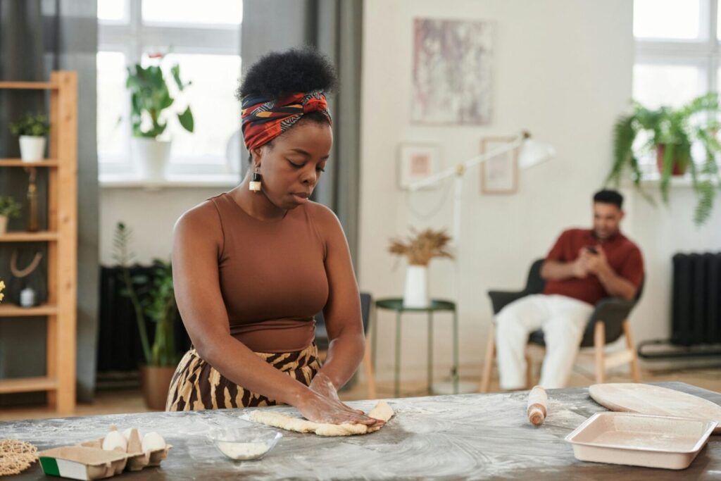 African woman with afro hair kneads dough on a wooden table in a cozy indoor setting with natural light.