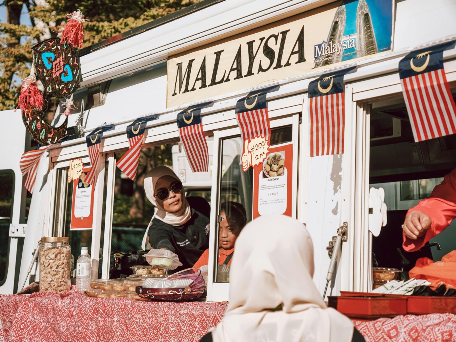 A vibrant Malaysian street food vendor serving customers at a market stall with traditional flags.