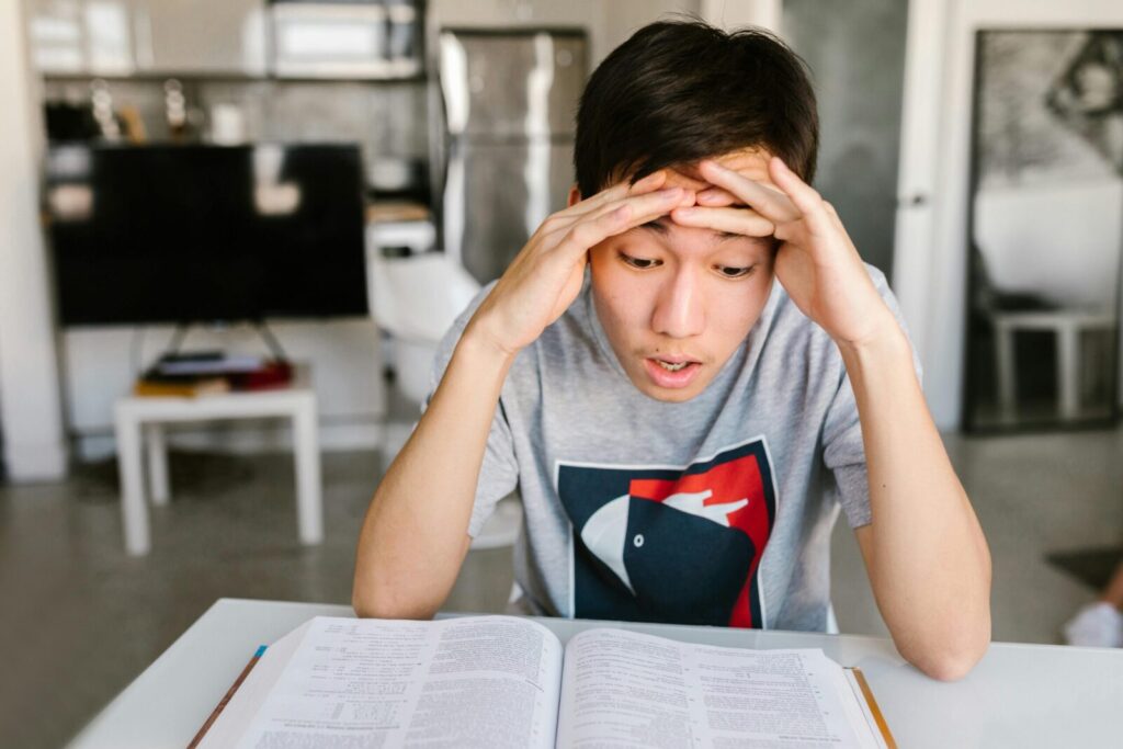 A young man studying at home, appearing stressed over his books at a table indoors.