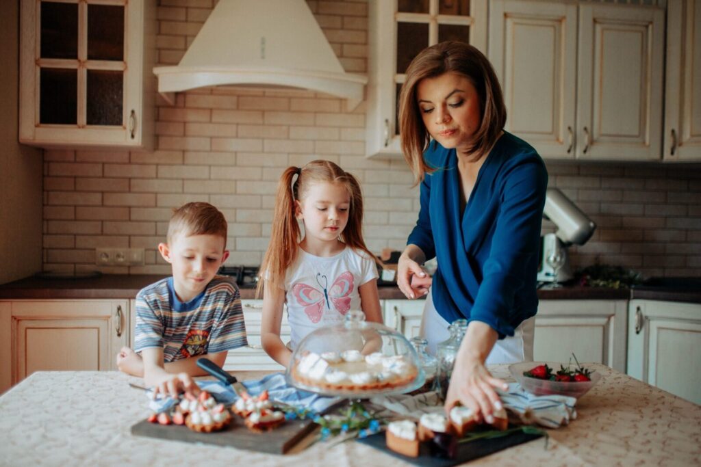 Children and lady in casual clothes standing near table with tasty desserts with cream and strawberry in light kitchen with cupboards