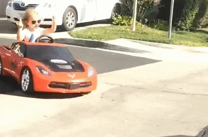 Video gif. Preschool age girl in sunglasses holds up both hands with 2 fingers up in a rock on gesture. She rides in a red toy sports car that turns and slides out into the street.
