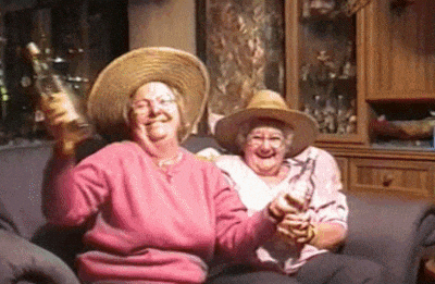 Video gif. Two senior women sit on a couch wearing straw hats, gleefully dancing around with their bottles of moonshine.