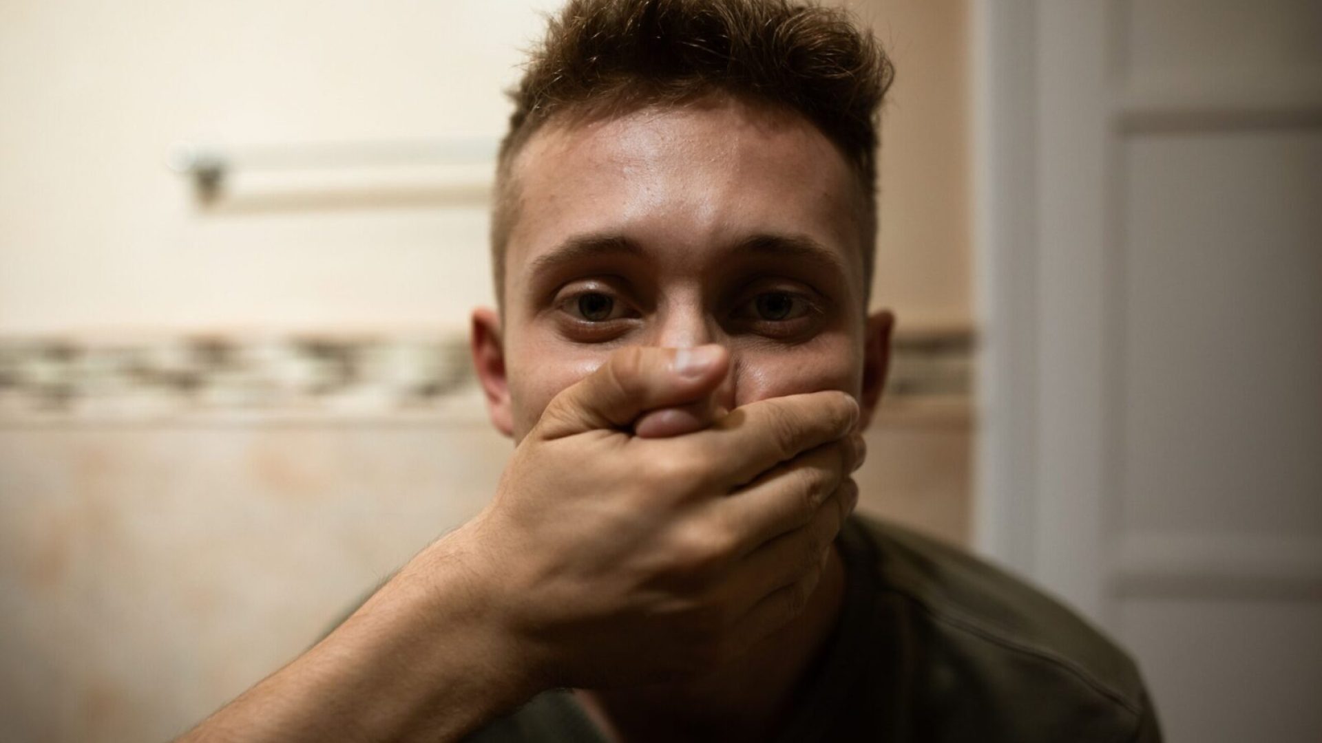 A young man indoors with a serious expression, hand covering his mouth, highlighting themes of silence and suppression.