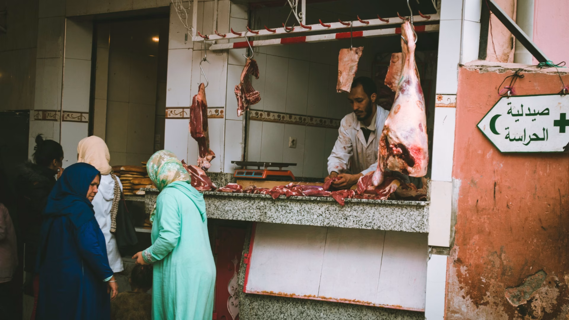 A lively outdoor market scene showing a butcher serving customers at a meat stall.